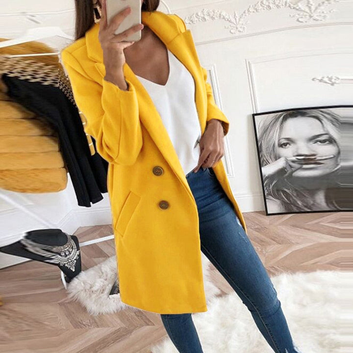 Frau in gelbem Mantel, weißem Top und blauer Jeans im modernen Wohnzimmer.