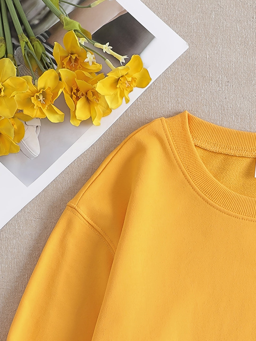 Gelbes Sweatshirt aus Baumwolle neben gelben Blumen auf beigem Hintergrund.