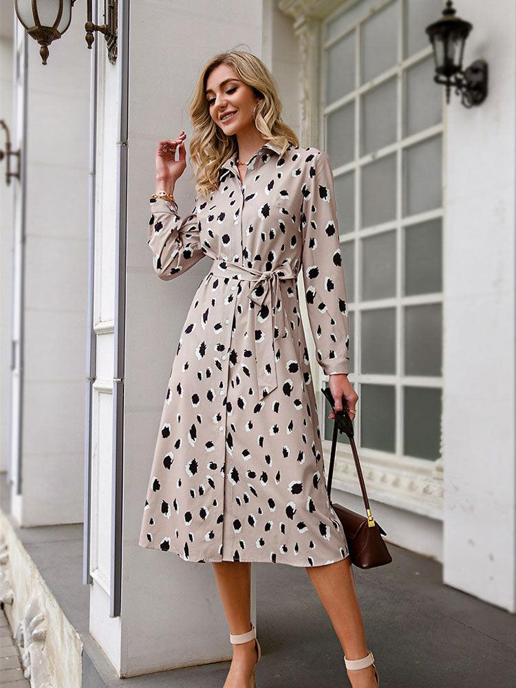 Frau in beigem, gepunktetem Hemdkleid mit Gürtel, steht vor einem Gebäude, modisch elegant.