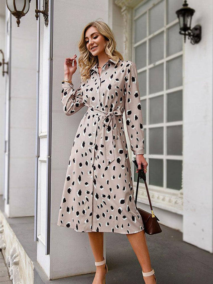 Frau in beigem, gepunktetem Hemdkleid mit Gürtel, steht vor einem Gebäude, modisch elegant.