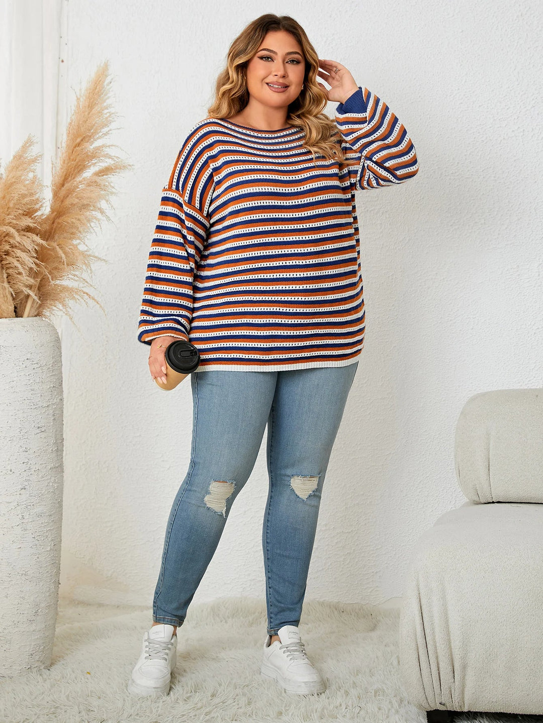 Frau in gestreiftem Pullover, blaue Jeans, weiße Sneaker, Kaffeebecher, gemütliches Wohnzimmer.