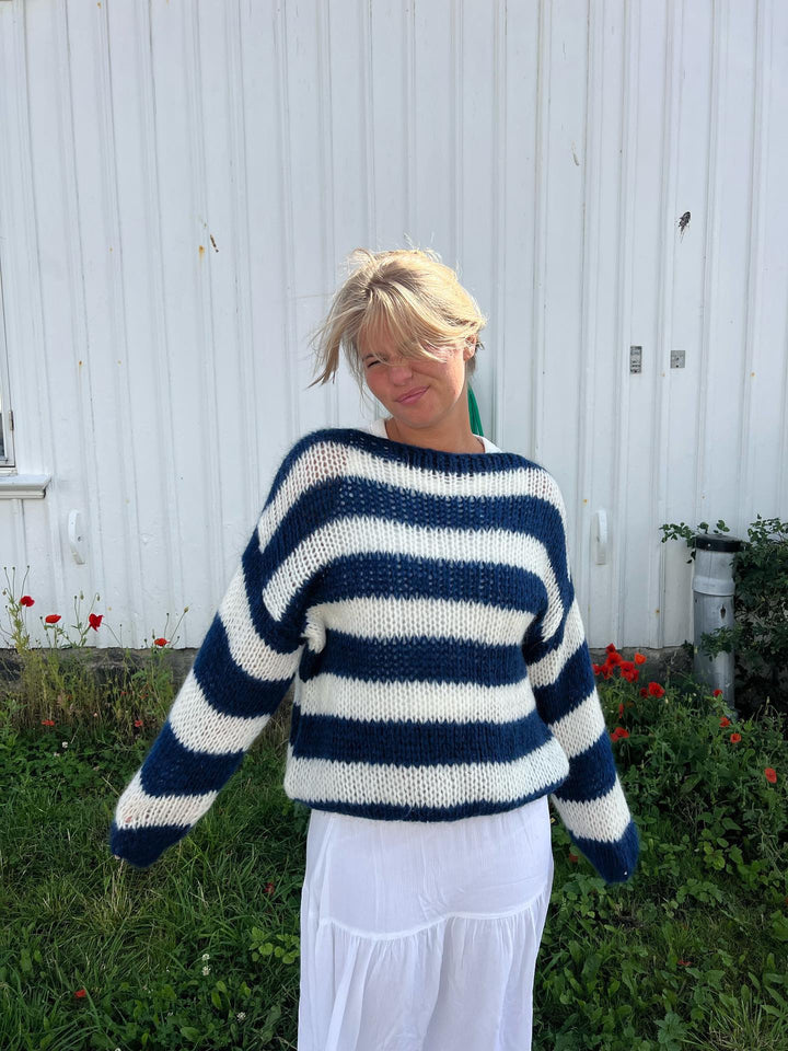 Frau in gestreiftem Pullover vor weißer Wand, blau-weiß, Strickmode, lässiger Stil.