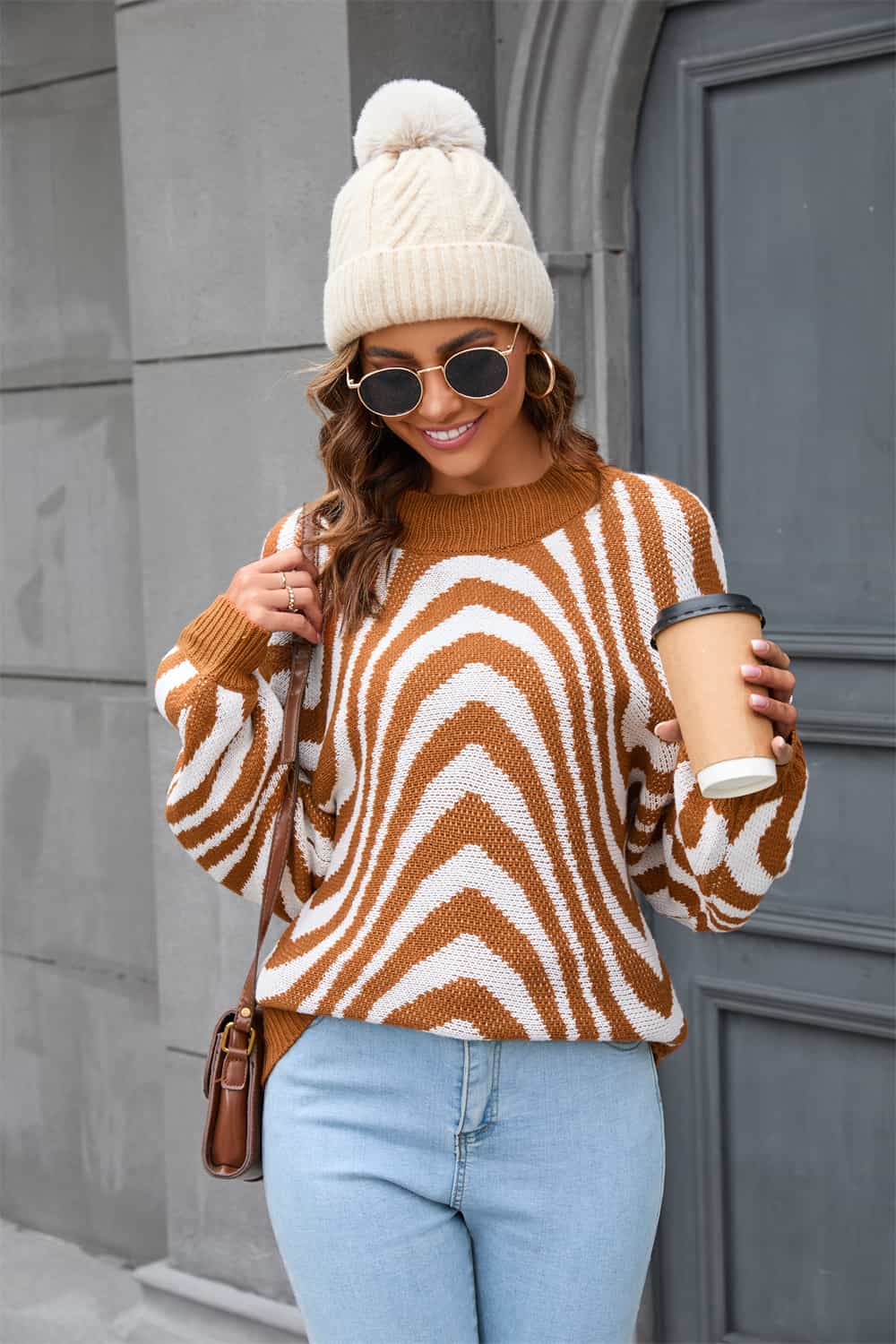 Frau in gestreiftem Pullover, blauer Jeans, weißer Mütze, Sonnenbrille, hält Kaffeebecher.