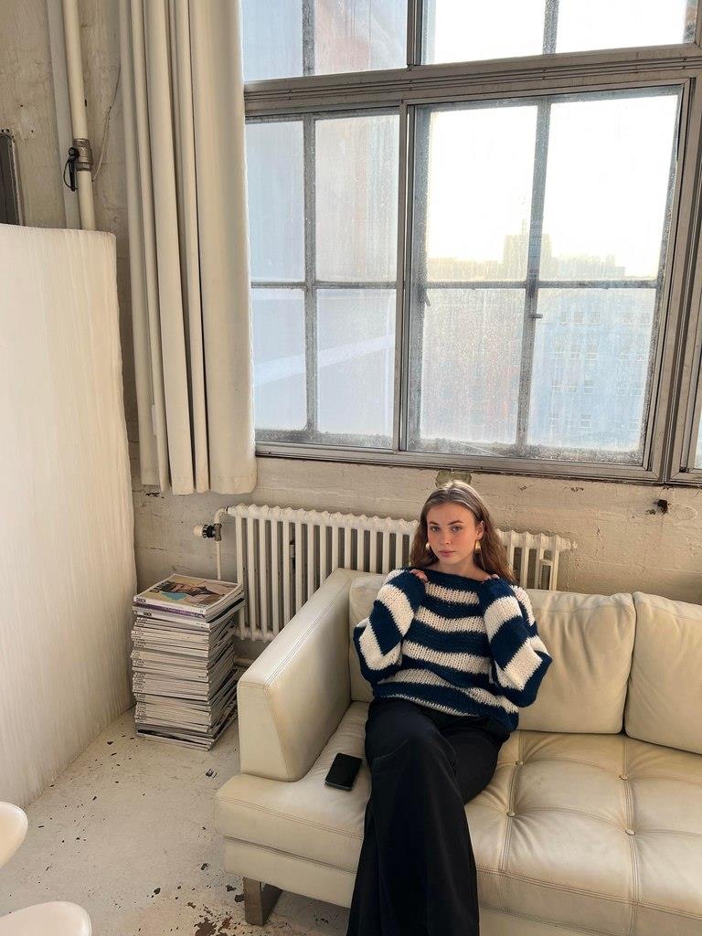 Frau in gestreiftem Pullover sitzt auf weißem Sofa, Loft-Wohnung, große Fenster, Tageslicht.