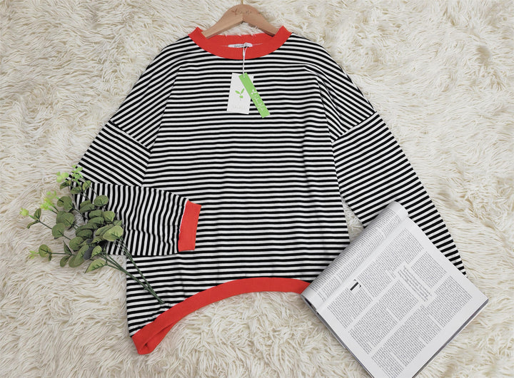 Schwarz-weiß gestreifter Pullover mit roten Bündchen auf Teppich, neben Zeitung und Pflanze.