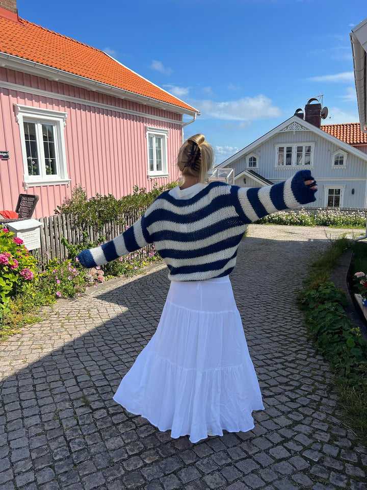 Frau im gestreiften Pullover und weißem Rock vor skandinavischem Haus, blauer Himmel.