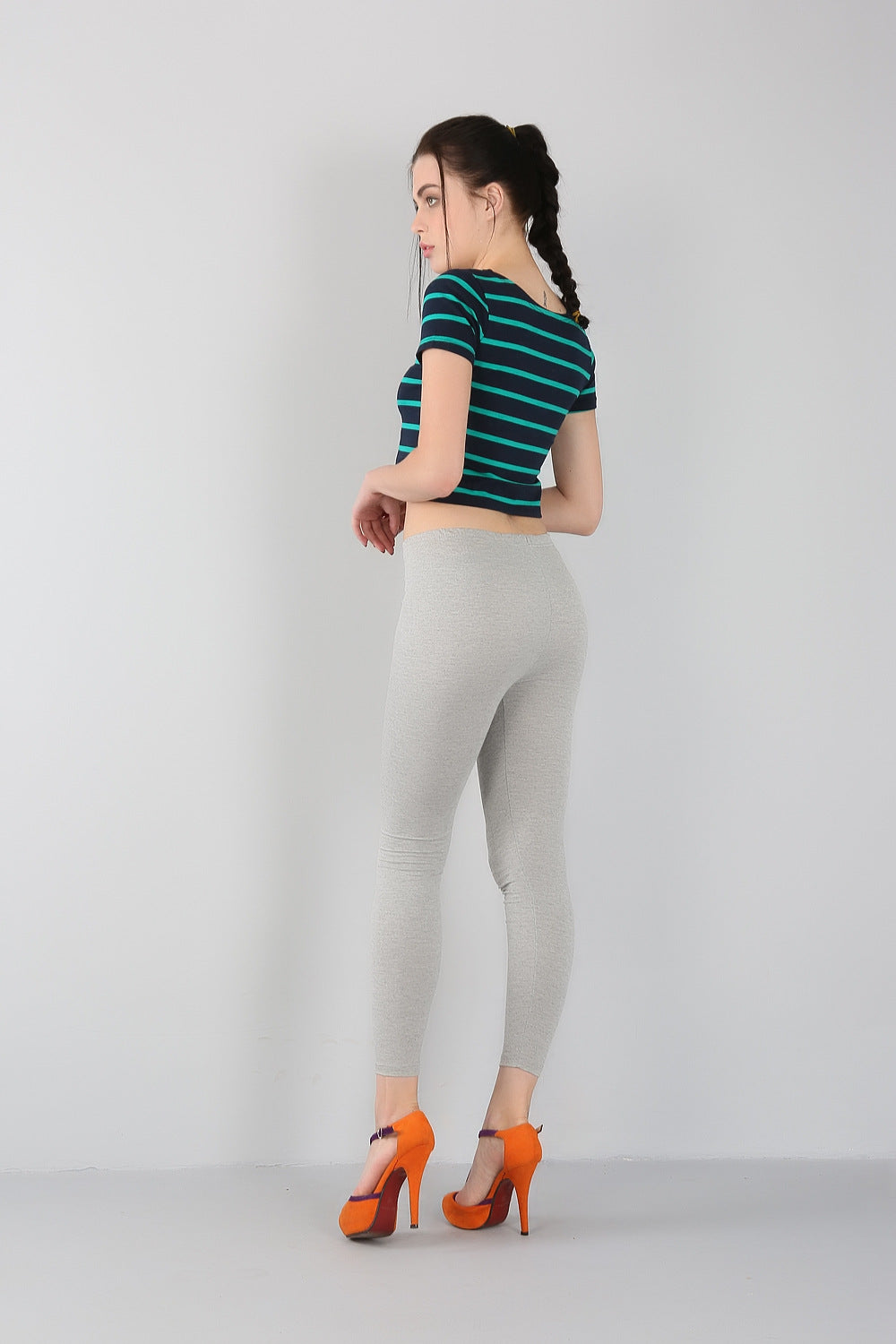 Frau in gestreiftem T-Shirt, grauen Leggings, orangefarbenen High Heels, seitlich stehend.