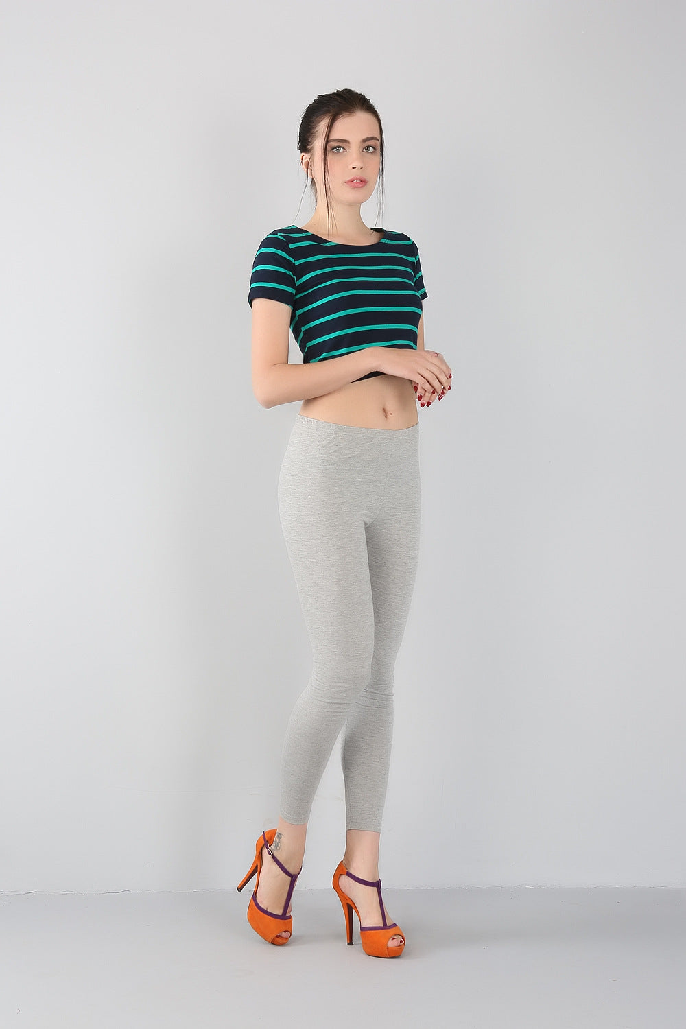 Frau in gestreiftem T-Shirt, grauer Leggings und orangefarbenen High Heels vor grauem Hintergrund.