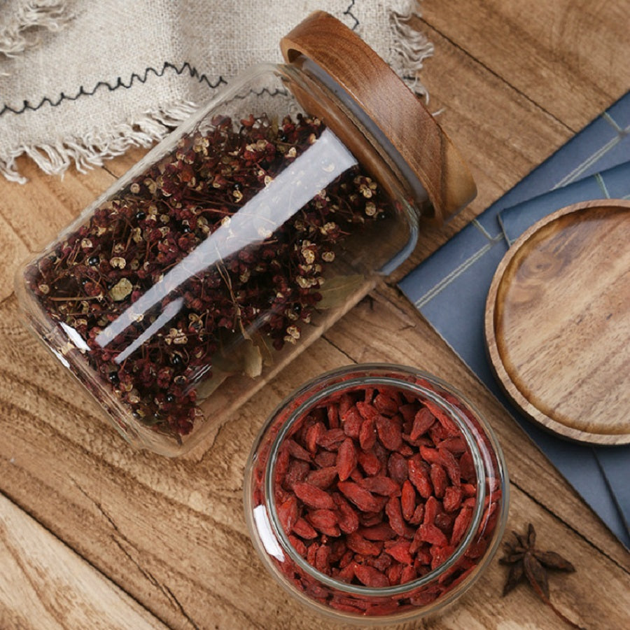 Glasbehälter mit Holzdeckel, gefüllt mit getrockneten Kräutern und Goji-Beeren auf Holztisch.