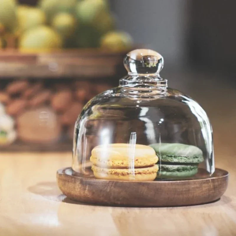 Zwei Macarons unter Glasglocke auf Holzteller, gelb und grün, auf Holztisch.