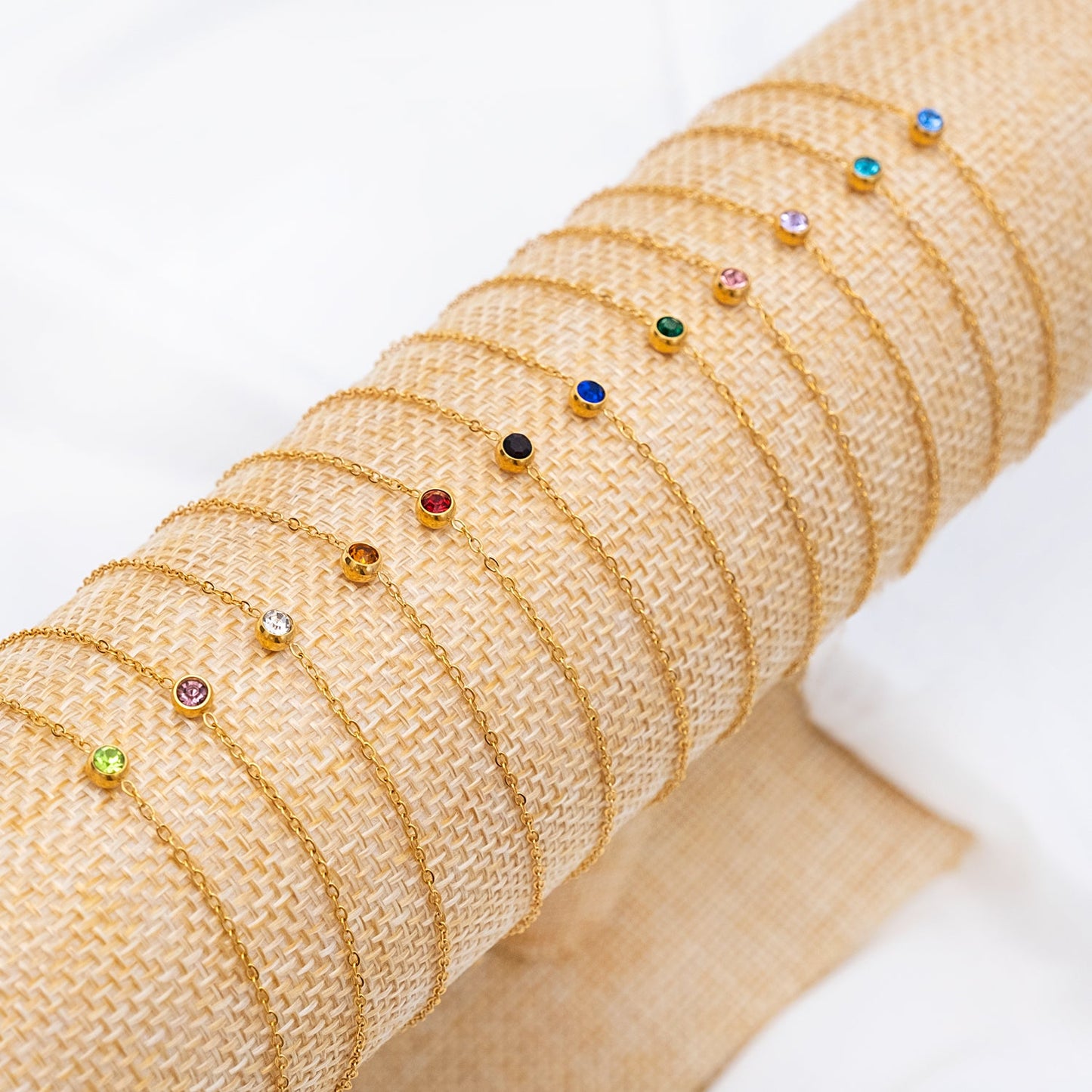Goldene Armbänder mit Geburtssteinen, verschiedene Farben, auf Jute-Hintergrund.
