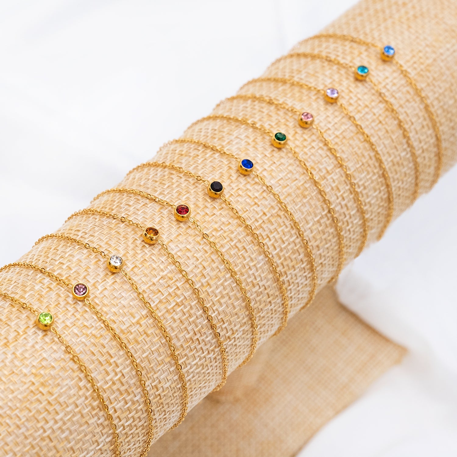 Goldene Armbänder mit Geburtssteinen, verschiedene Farben, auf Jute-Hintergrund.
