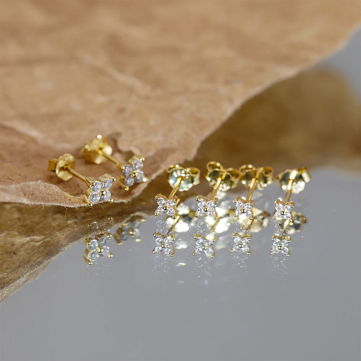 Goldene Ohrringe mit Zirkonia-Steinen, Blumenform, reflektiert auf glänzender Oberfläche.