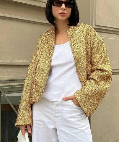Frau in goldener Paillettenjacke, weißem Top und Hose, Sonnenbrille, modisch, urbaner Stil.