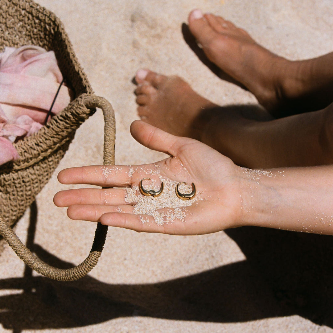 Goldene ovale Ohrringe auf Hand im Sand, Strandtasche, Sommeraccessoire.
