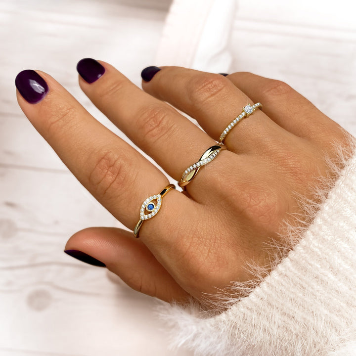 Damenhand mit drei goldenen Ringen, blauer Stein, Zirkonia, lila Nagellack, weißer Pullover.
