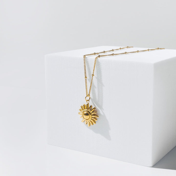 Goldene Sonnen-Halskette auf weißem Würfel, elegantes Schmuckdesign, modisches Accessoire.