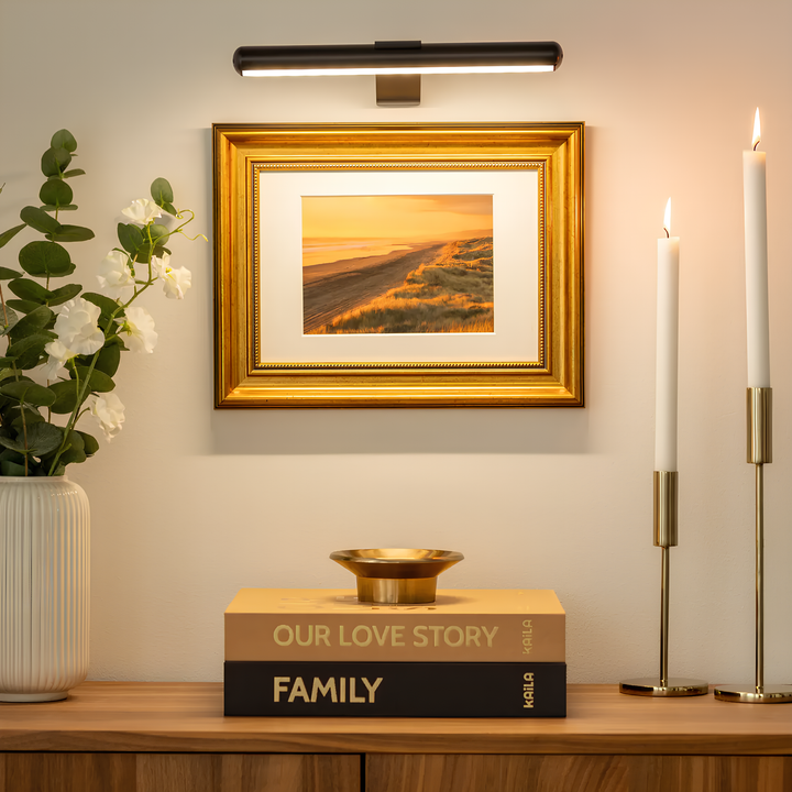 Goldener Bilderrahmen mit Landschaftsfoto, umgeben von Kerzen und Blumen, auf Holzkommode.