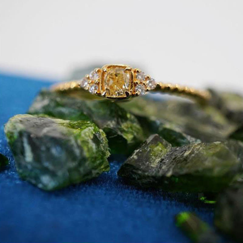 Goldener Diamantring mit gelbem Stein, umgeben von grünen Edelsteinen auf blauem Hintergrund.