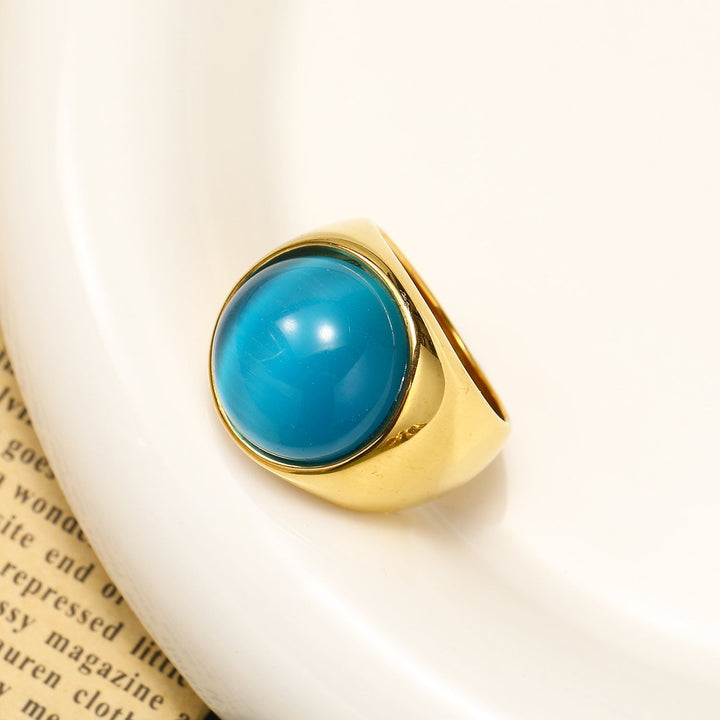 Goldener Ring mit großem, rundem, blauem Stein auf weißem Hintergrund, elegantes Schmuckdesign.