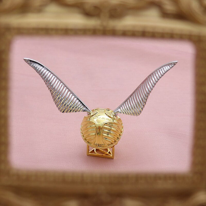 Goldener Schnatz Replik mit silbernen Flügeln, Harry Potter Sammlerstück, auf rosa Hintergrund.
