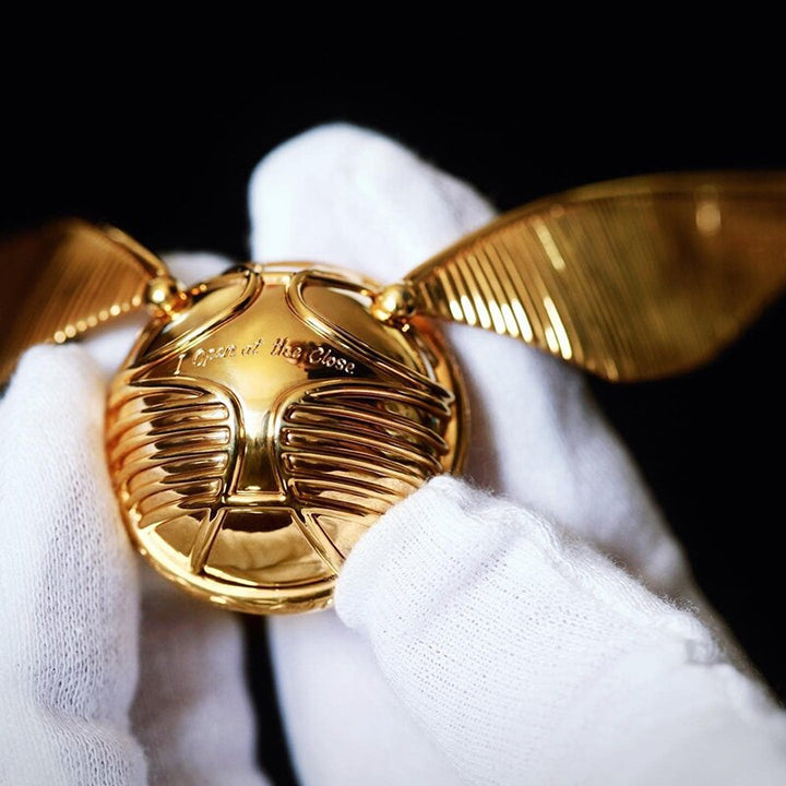 Goldener Schnatz Replik, glänzend, mit Flügeln, in weiß behandschuhten Händen gehalten.
