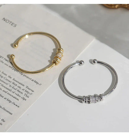 Zwei offene Armreifen, gold und silber, mit Strasssteinen auf Buchseite liegend.