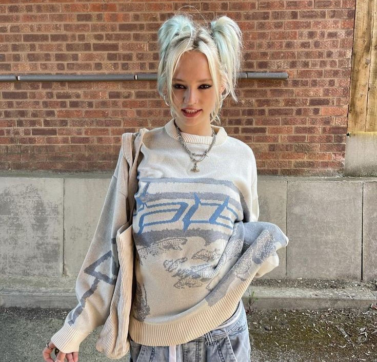 Frau mit blauem Pullover und Zöpfen vor Backsteinwand, lässiger Streetwear-Look.