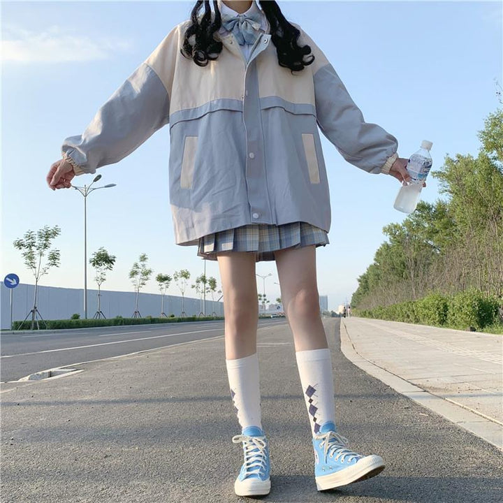 Frau in grauer Jacke und kariertem Rock, blaue Sneakers, steht auf Straße, hält Wasserflasche.