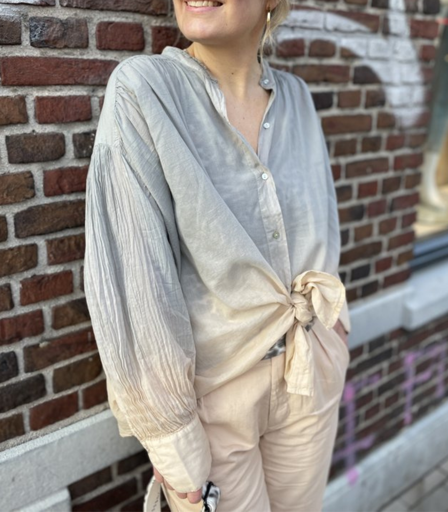 Frau in grauer Bluse mit Knoten, Stehkragen, langen Ärmeln, vor Ziegelwand.