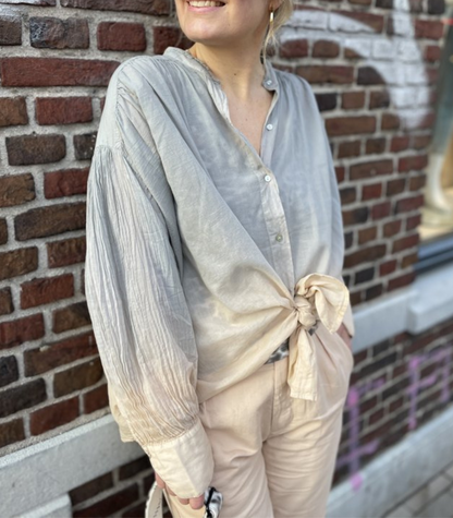 Frau in grauer Bluse mit Knoten, Stehkragen, langen Ärmeln, vor Ziegelwand.