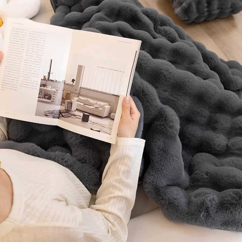 Person liest auf grauer, flauschiger Decke; gemütliche Wohnkultur, Entspannung, modernes Design.