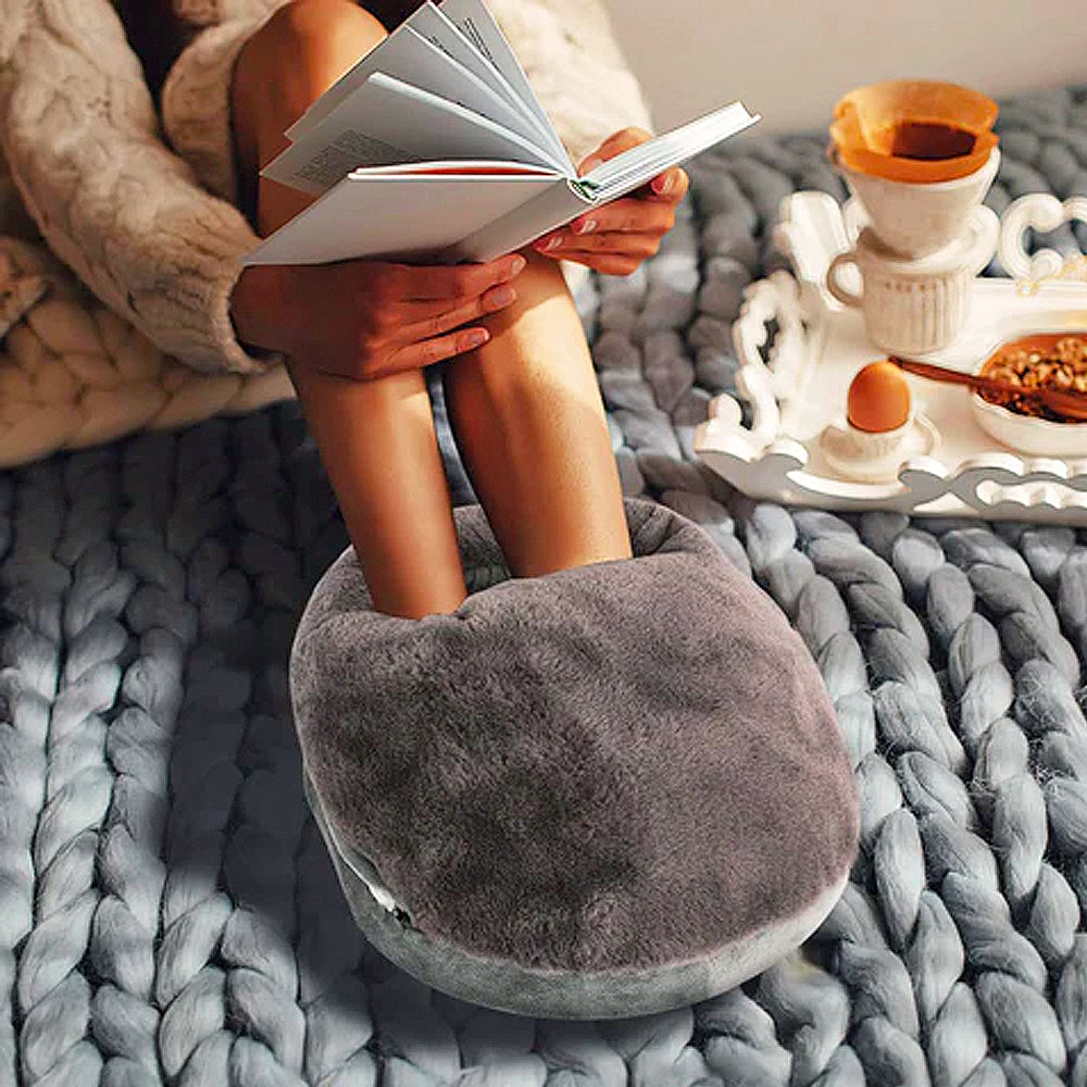 Flauschiger grauer Fußwärmer Pantoffel auf gestrickter Decke, gemütliches Lesen im Bett.