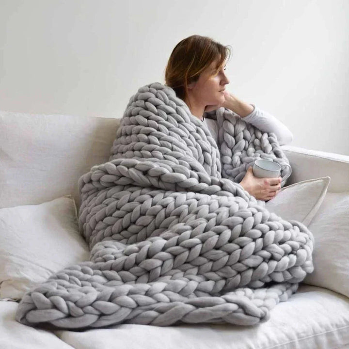 Frau mit grauer, grob gestrickter Decke auf Sofa, gemütlich, Tasse haltend, Wohnzimmer.