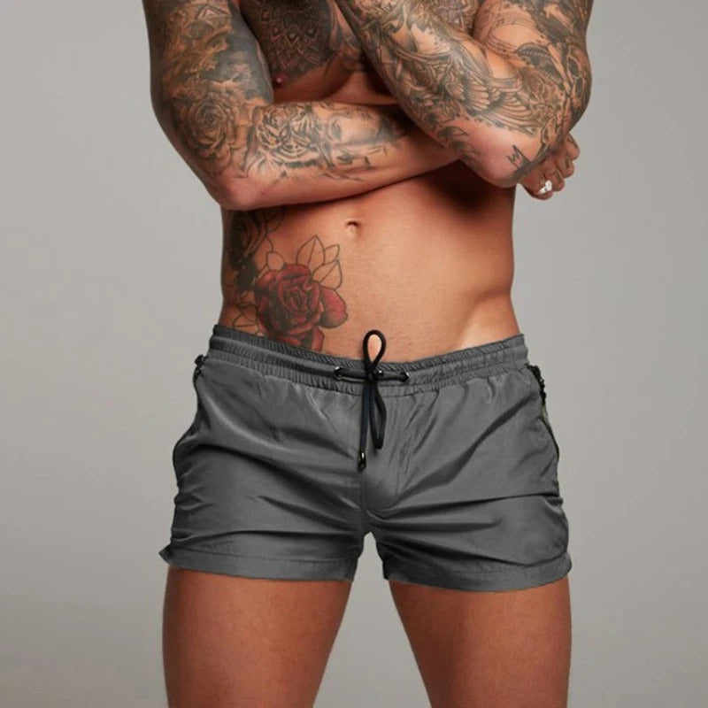 Mann mit tätowiertem Oberkörper trägt graue Badehose, sportlich, modern, Strandmode.