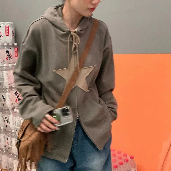 Frau in grauem Kapuzenpullover mit Stern, braune Umhängetasche, Jeans, Getränkeflaschen im Hintergrund.