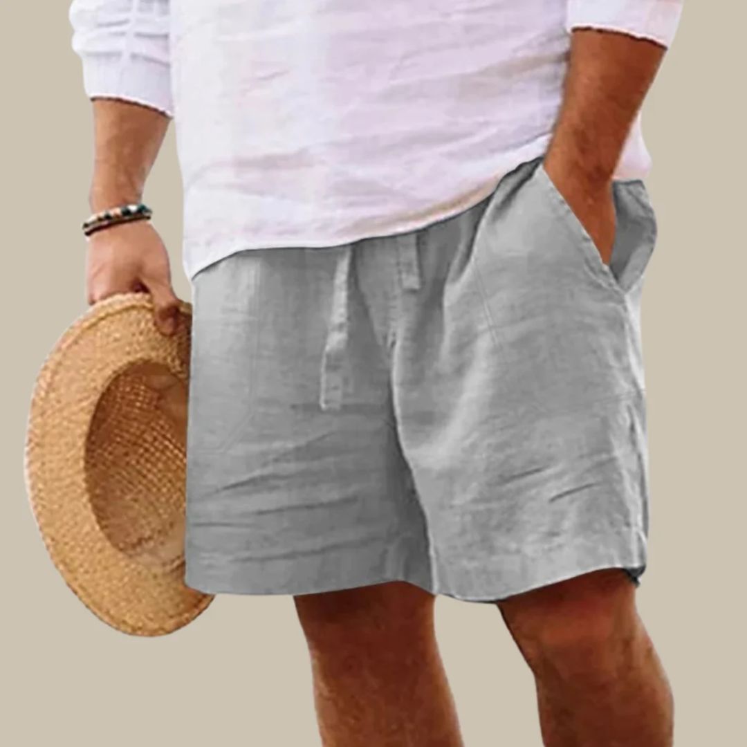 Mann in grauen Leinen-Shorts und weißem Hemd hält Strohhut, Sommermode.