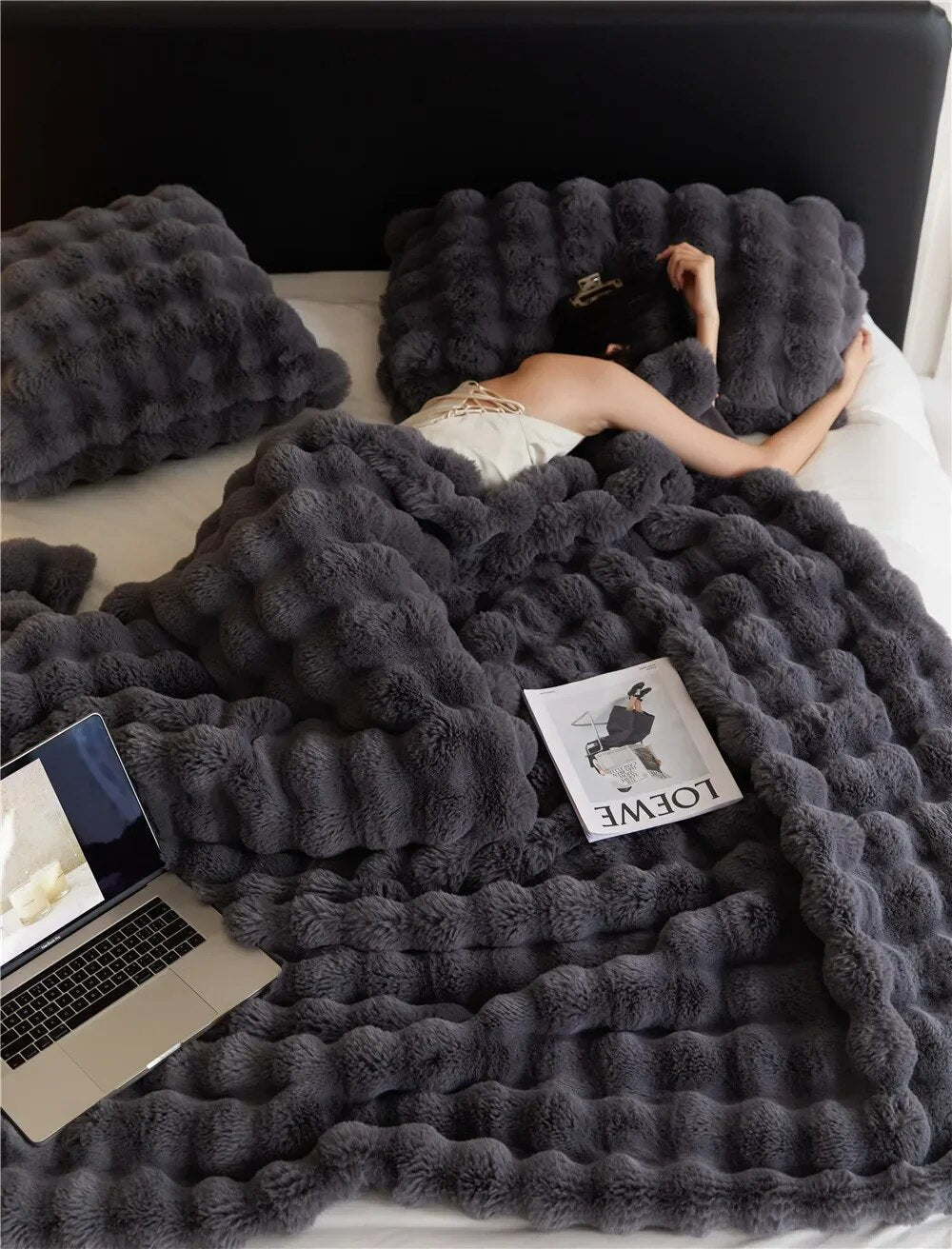 Graue, flauschige Bettdecke und Kissen auf Bett, Person schläft, Laptop und Magazin daneben.