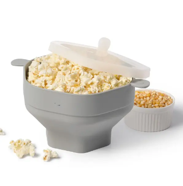 Graue Silikon-Popcornschüssel mit Deckel, gefüllt mit frischem Popcorn, daneben Maiskörner.