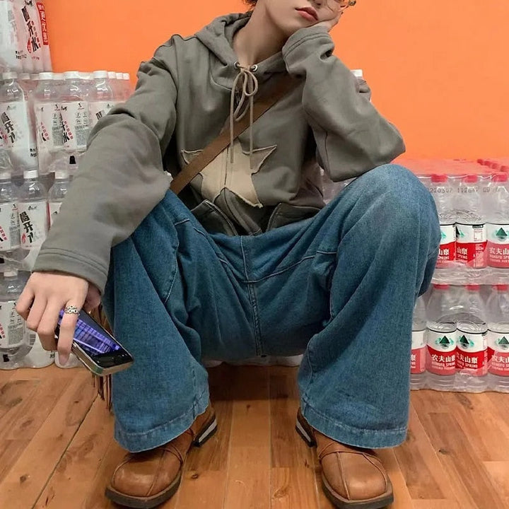 Person in grauem Hoodie und blauer Jeans sitzt vor Wasserflaschen, hält Smartphone, trägt braune Schuhe.