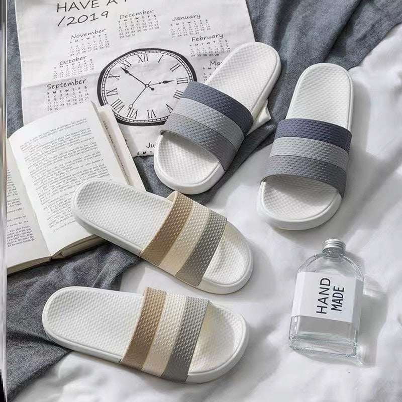Vier Paar Badeschlappen, grau und beige, auf Bett mit Buch und Uhr, modernes Design.