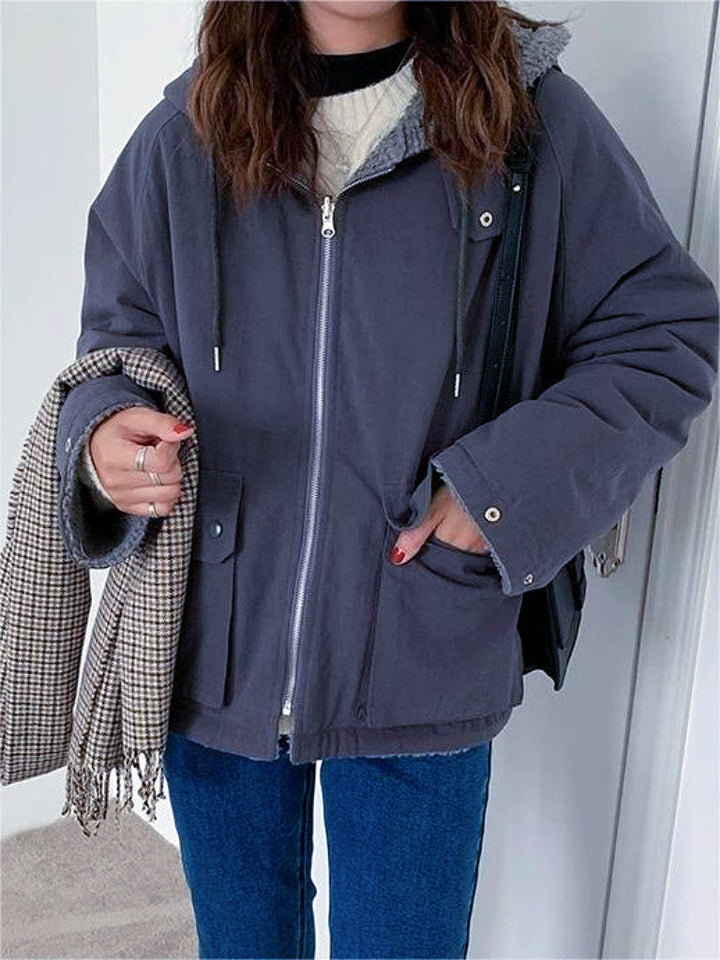 Frau in grauer Winterjacke mit Kapuze, hält karierten Schal, trägt blaue Jeans.