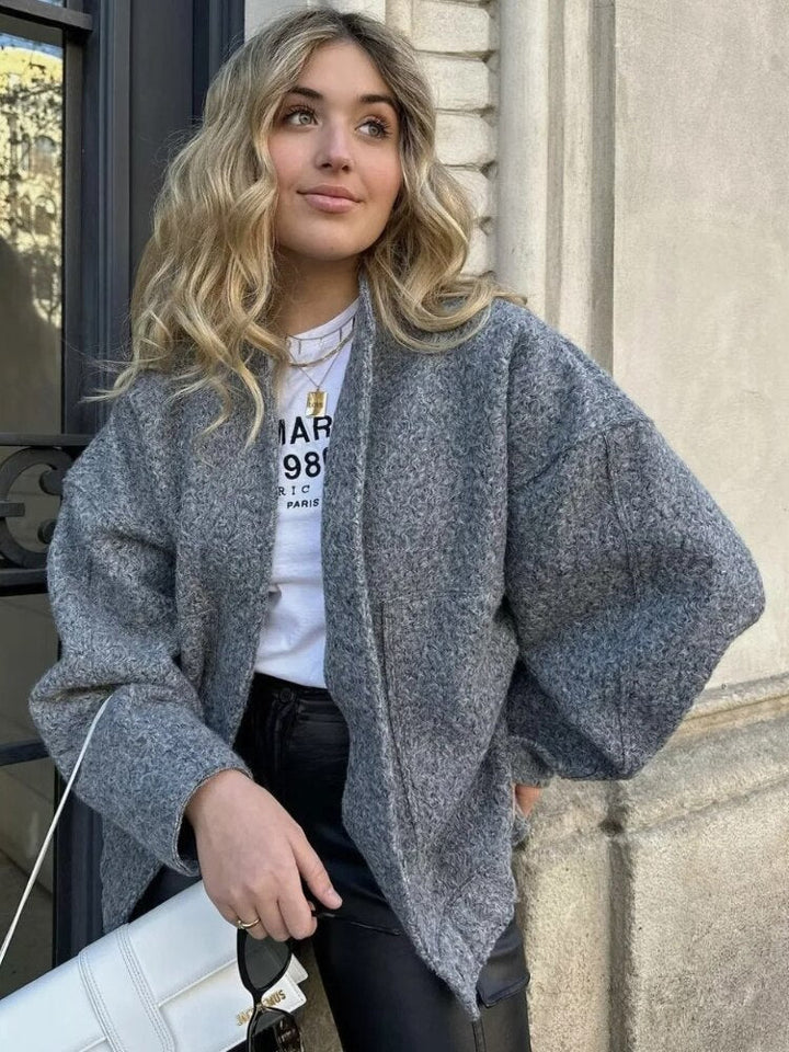 Frau in grauem Wollmantel, weißem T-Shirt, schwarzer Lederhose, hält weiße Handtasche.