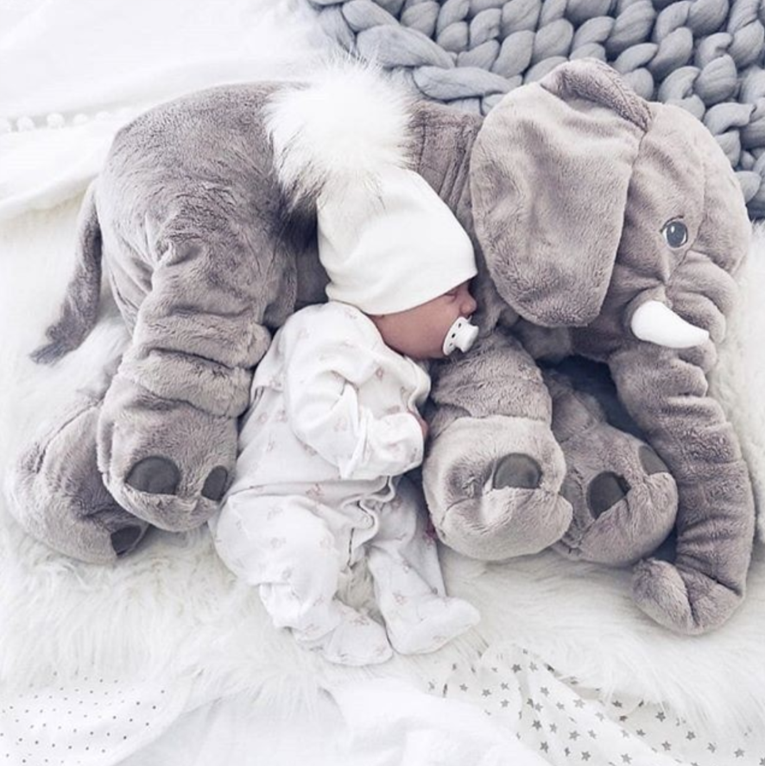 Baby mit grauem Plüschelefanten, weißer Mütze und Schnuller auf weicher Decke.