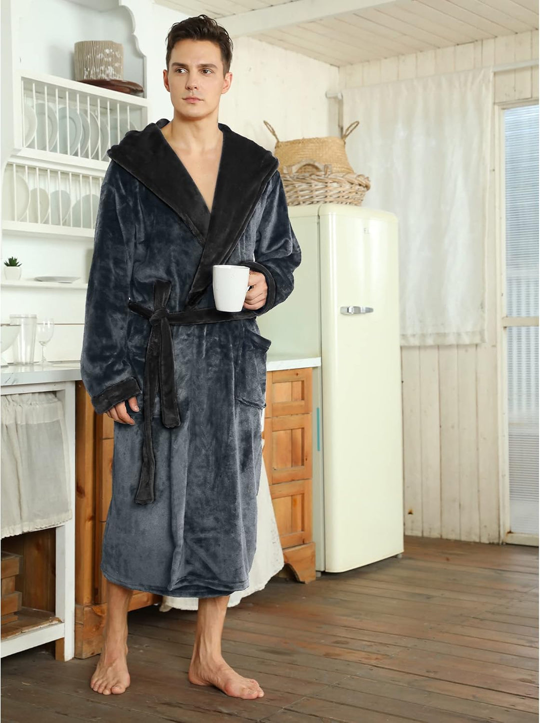 Mann im grauen Fleece-Bademantel mit Kapuze, hält Tasse, steht in rustikaler Küche.