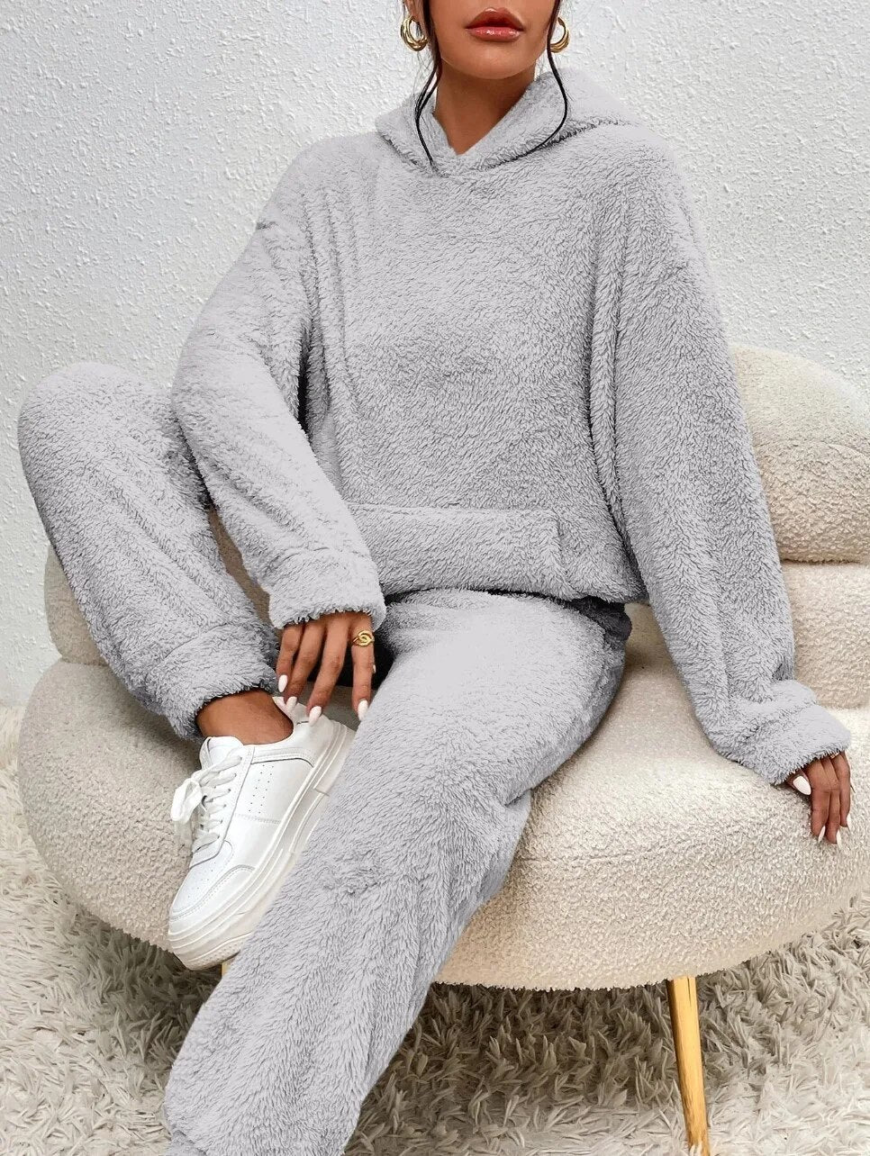 Frau in grauem Fleece-Hoodie und Jogginghose auf Sessel, weiße Sneaker, gemütliche Mode.
