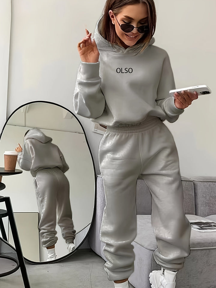 Frau in grauem Leder-Trainingsanzug mit Smartphone und Kaffeetasse, Spiegel im Hintergrund.