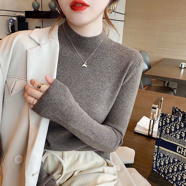 Frau in grauem Rollkragenpullover und weißem Blazer, trägt Kette, sitzt in modernem Café.