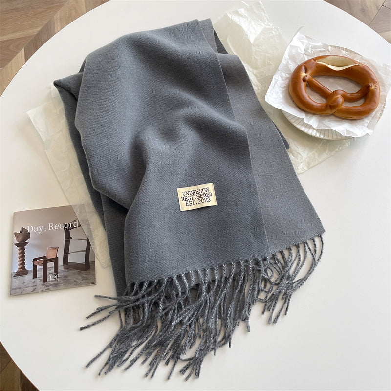 Grauer Wollschal mit Fransen auf Tisch neben Brezel und Magazin, stilvolles Accessoire.