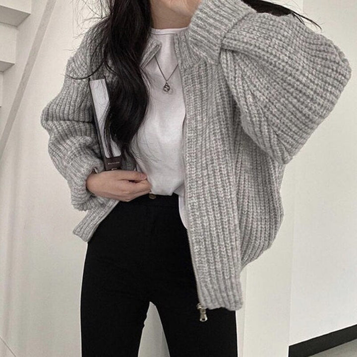 Frau in grauer Strickjacke, weißem T-Shirt, schwarzer Hose, hält Buch, modisch, lässig.